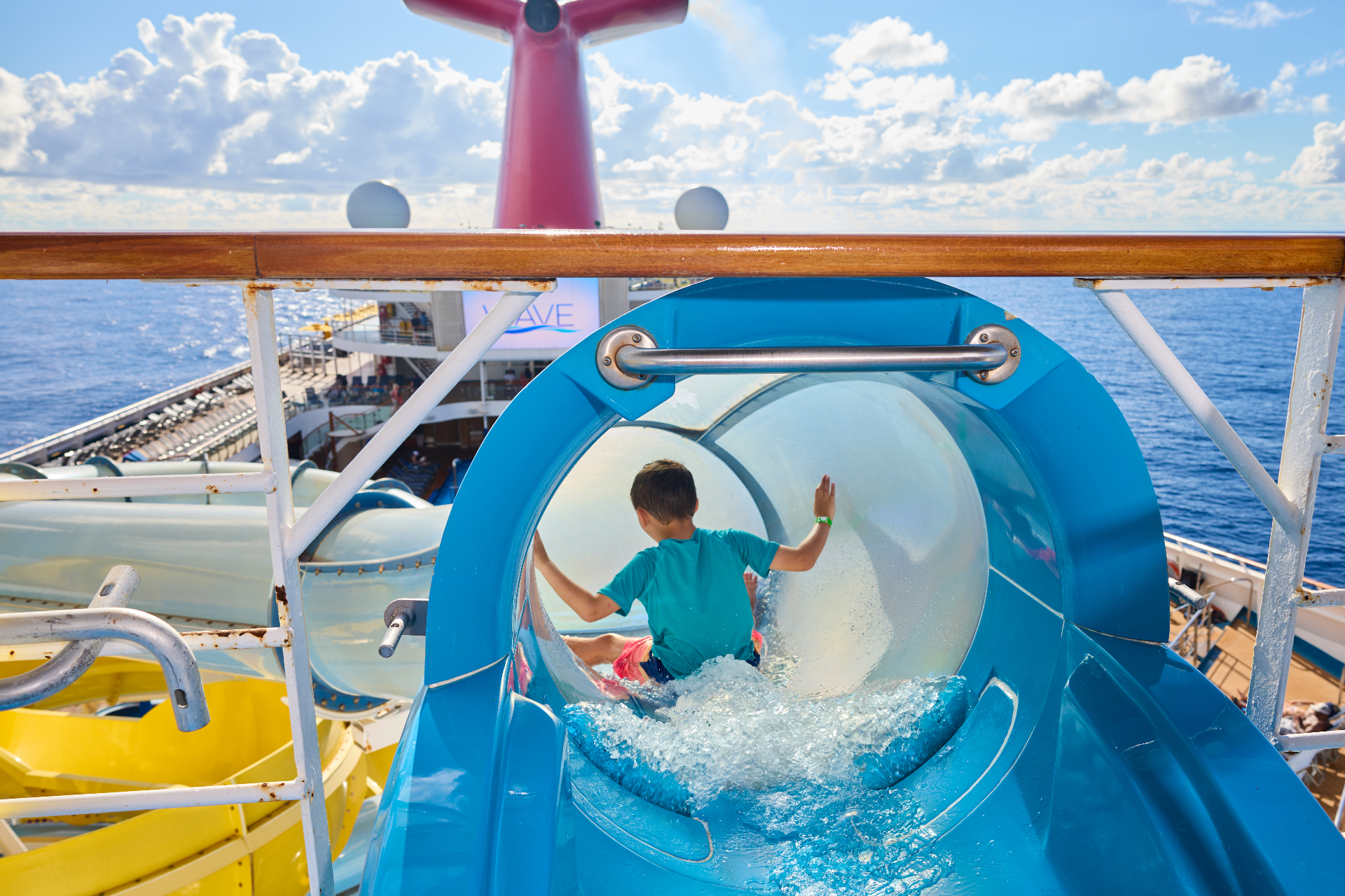 A child riding the blue tube waterslide on a Carnival ship deck with the ocean behind